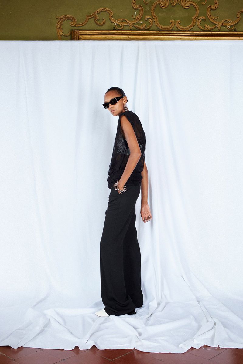 A tall woman in a sheer black top, sunglasses, and black palazzo pants stands on draped white fabric in front of a gold-trimmed wall.