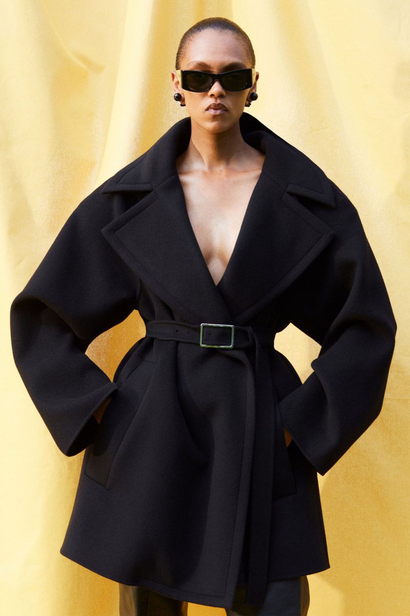 A model posing in a black belted coat, rectangular sunglasses, and black earrings against a textured yellow backdrop, fashion forward.