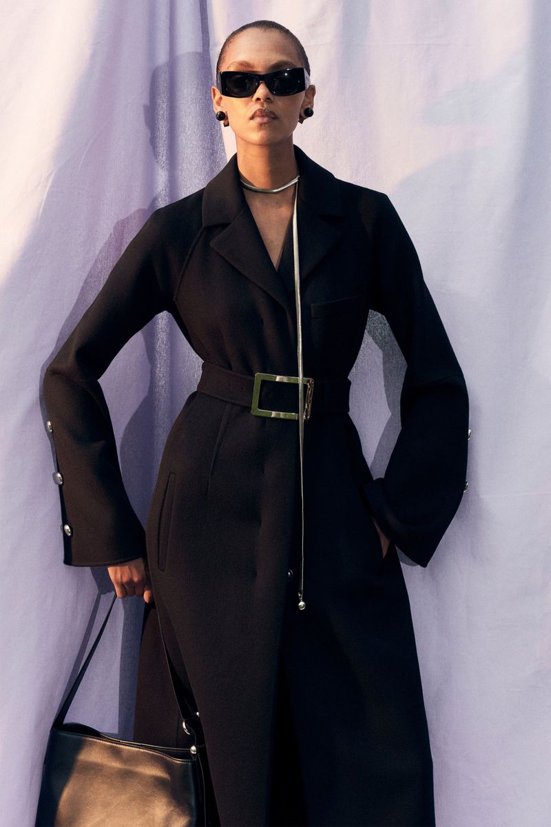 Elegant adult female poses in a belted black coat with wide sleeves, accessorized with shades, necklace and a purse against a soft purple backdrop.