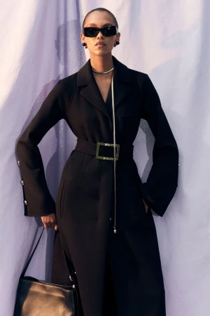 Elegant adult female poses in a belted black coat with wide sleeves, accessorized with shades, necklace and a purse against a soft purple backdrop.