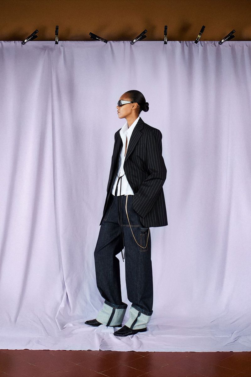 A lone woman with a sleek bun is styled in a pinstripe blazer and dark denim pants, posing against a lavender curtain backdrop.