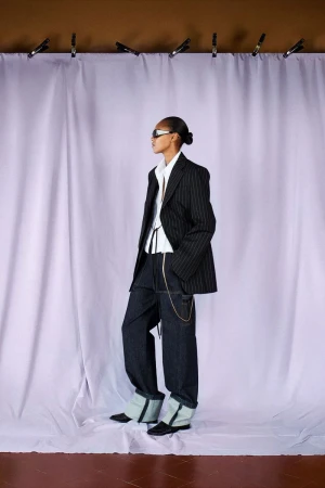 A lone woman with a sleek bun is styled in a pinstripe blazer and dark denim pants, posing against a lavender curtain backdrop.