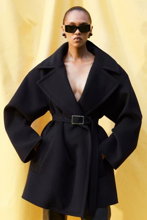 Model wearing a black belted coat and black sunglasses against a yellow draped background for Sportmax Resort 2026