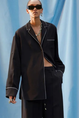 Model in black pajama-style shirt with white piping and sunglasses standing against blue draped fabric backdrop