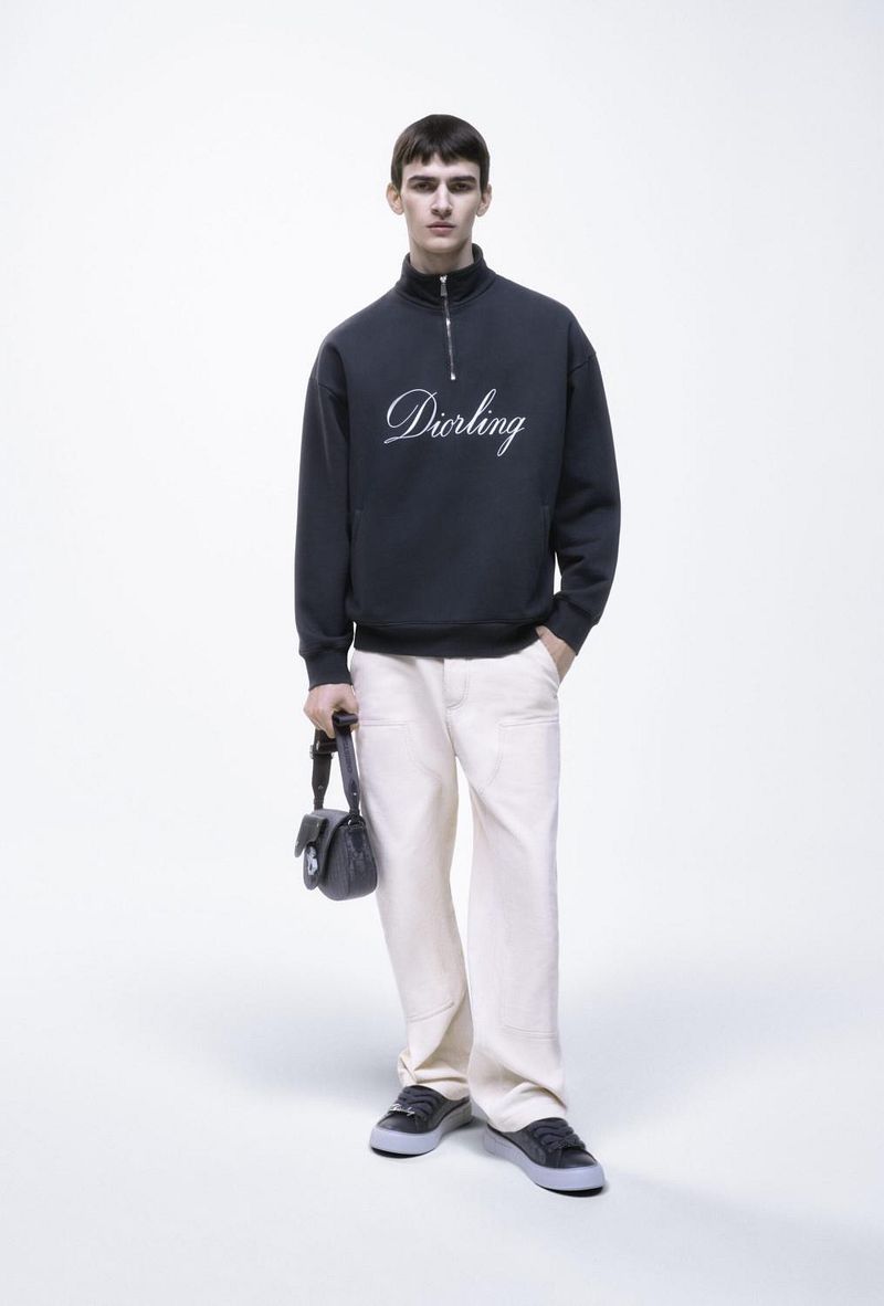 Male model poses in a studio wearing a dark-blue sweatshirt with 'Darling' embroidered, light-beige pants, black shoes, and a black purse.