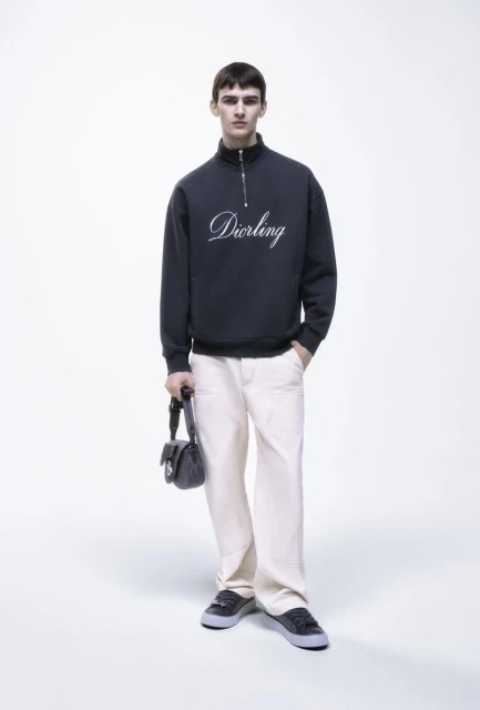 Male model poses in a studio wearing a dark-blue sweatshirt with 'Darling' embroidered, light-beige pants, black shoes, and a black purse.