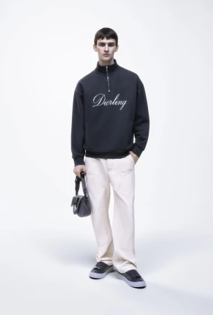 Male model poses in a studio wearing a dark-blue sweatshirt with 'Darling' embroidered, light-beige pants, black shoes, and a black purse.