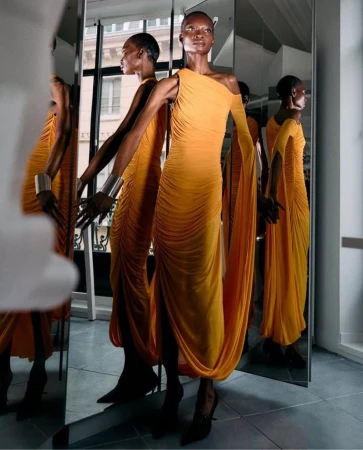 Woman in orange gown poses in photo studio with mirrors, showcasing fashion, style, and elegance in bright, stylized setting.