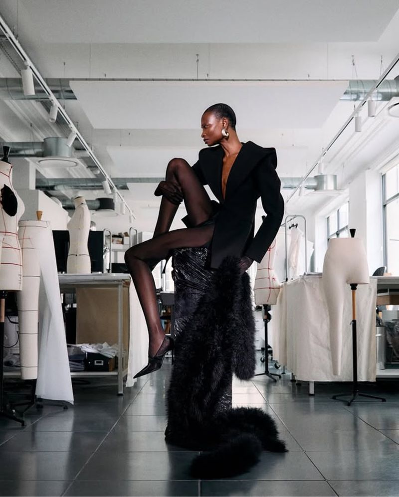 Fashion editorial shot of an African American woman in a black blazer and tights, posing on a metallic sculpture in a studio.