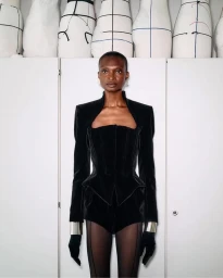 Model Debra Shaw in a black velvet Mugler jacket and corset standing in front of atelier dressmaker forms.