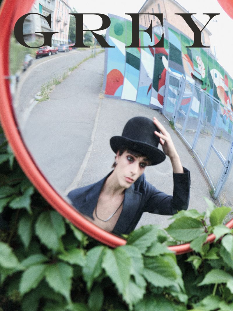 Mirror selfie of a young adult wearing a black bowler hat and a dark jacket, with green plants surrounding the mirror and a cityscape backdrop.
