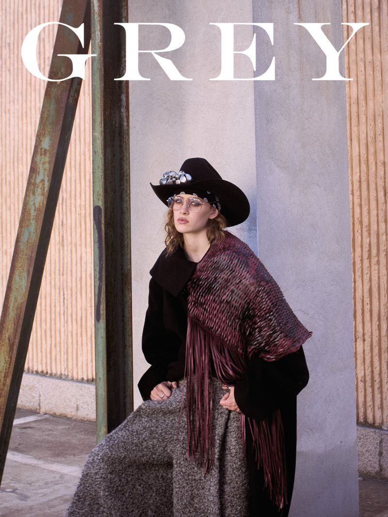 Fashion editorial shot of woman in black coat, hat, and grey skirt. She has glasses and poses in an outdoor environment with neutral colours.