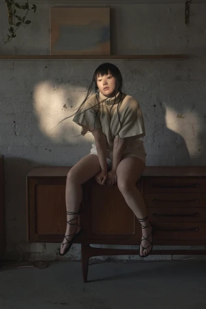 A young woman with dark hair poses while sitting on a vintage cabinet in a minimalist interior with neutral colors and diffused natural light.