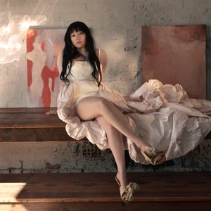 Woman with long dark hair wearing a white dress and beige heeled sandals seated on a wooden ledge against a textured wall with artwork.