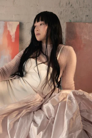 Yerin Ha seated wearing a pink ribbed knit top and white dress against a textured wall with artwork by Russh Magazine