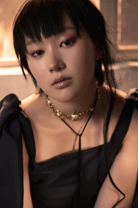 Portrait of a young woman with dark hair wearing a black dress and a pearl and gold choker necklace