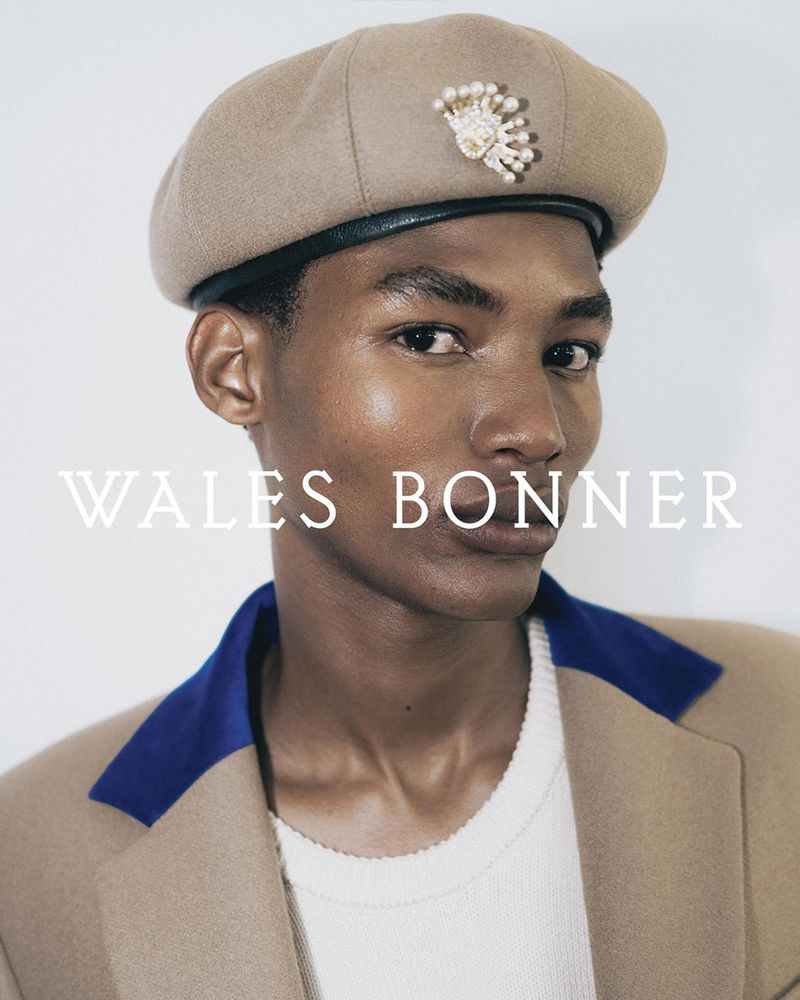 Close-up studio shot of a Black male model wearing a beige beret with jeweled detail, a white sweater, and a beige coat with blue trim.
