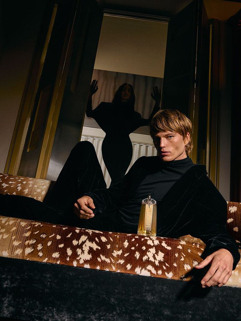 Male model reclines on a patterned couch next to a gold perfume bottle. A female figure stands silhouetted in the background doorway.