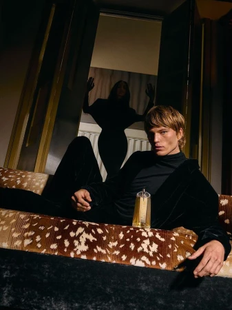 Male model reclines on a patterned couch next to a gold perfume bottle. A female figure stands silhouetted in the background doorway.
