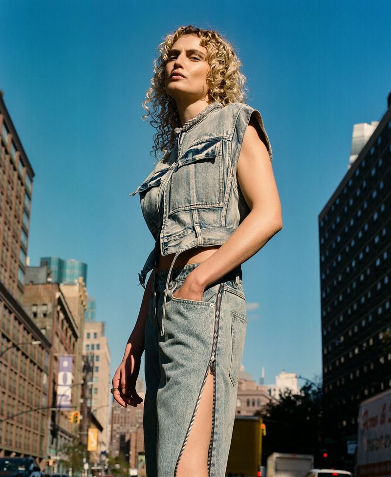Fashion model in a denim vest and skirt poses on a city street on a sunny day with buildings and blue sky in the background.