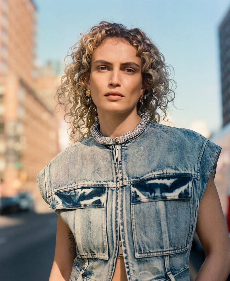 Adult woman with curly blonde hair poses in a light blue denim vest with a white top and silver necklace in an urban environment.
