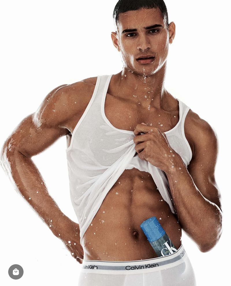 Male fashion model poses with a bottle of CK fragrance, wearing a white tank top and briefs against a bright, clean backdrop.
