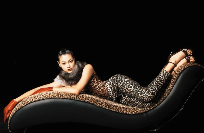 Adult female in animal-print attire poses on a black chaise lounge with a matching animal-print throw against a dark backdrop.