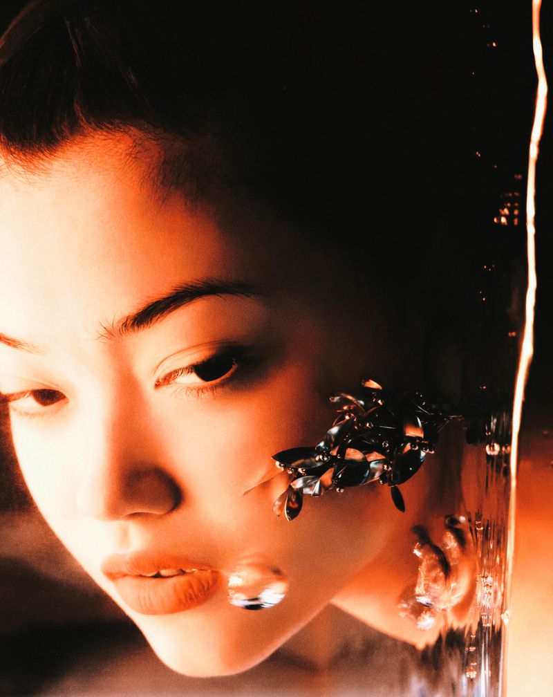 Close-up studio shot of a woman with dark hair and elaborate earrings, framed by flowing water and air bubbles against a dark background.