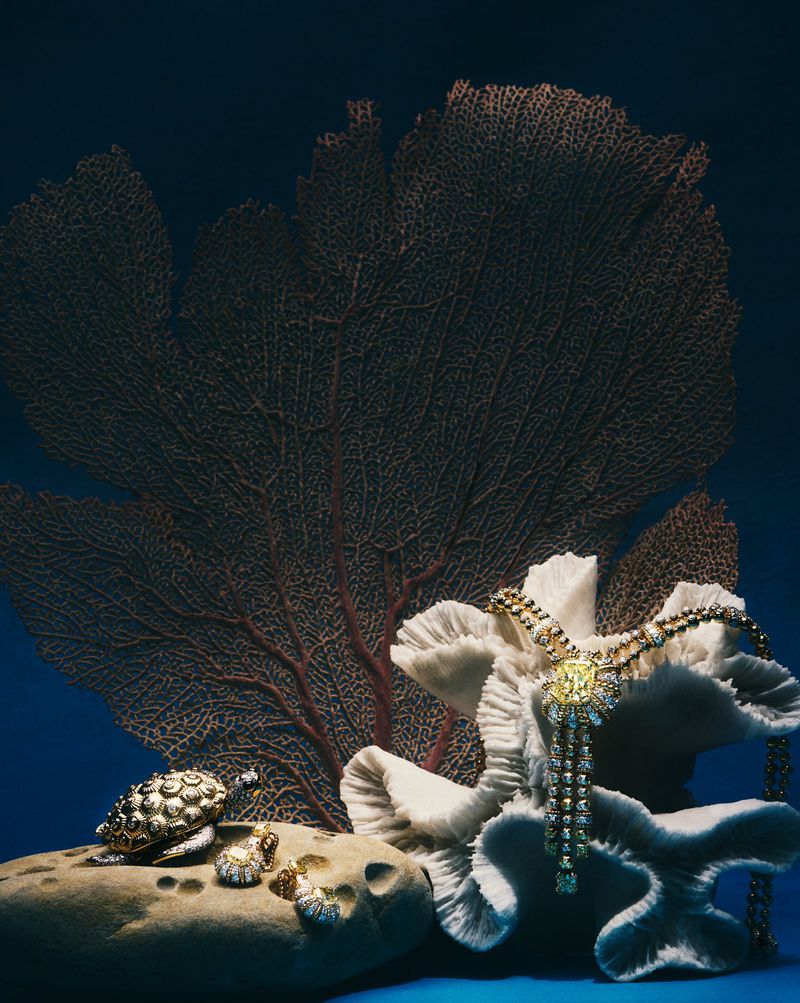 Still life shot of a gold and diamond necklace draped over white coral with gold turtles on a rock against a blue background.