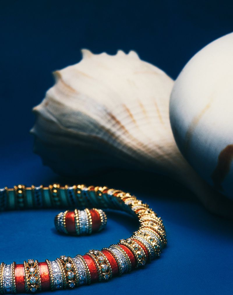 Gold luxury necklace and matching ring with red enamel and pavé stones beside large seashells on a deep blue surface.
