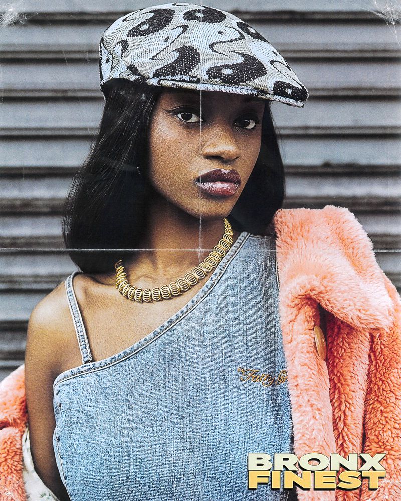 Portrait of a young Black woman wearing a patterned hat, gold necklace, and denim top against a roller door background.