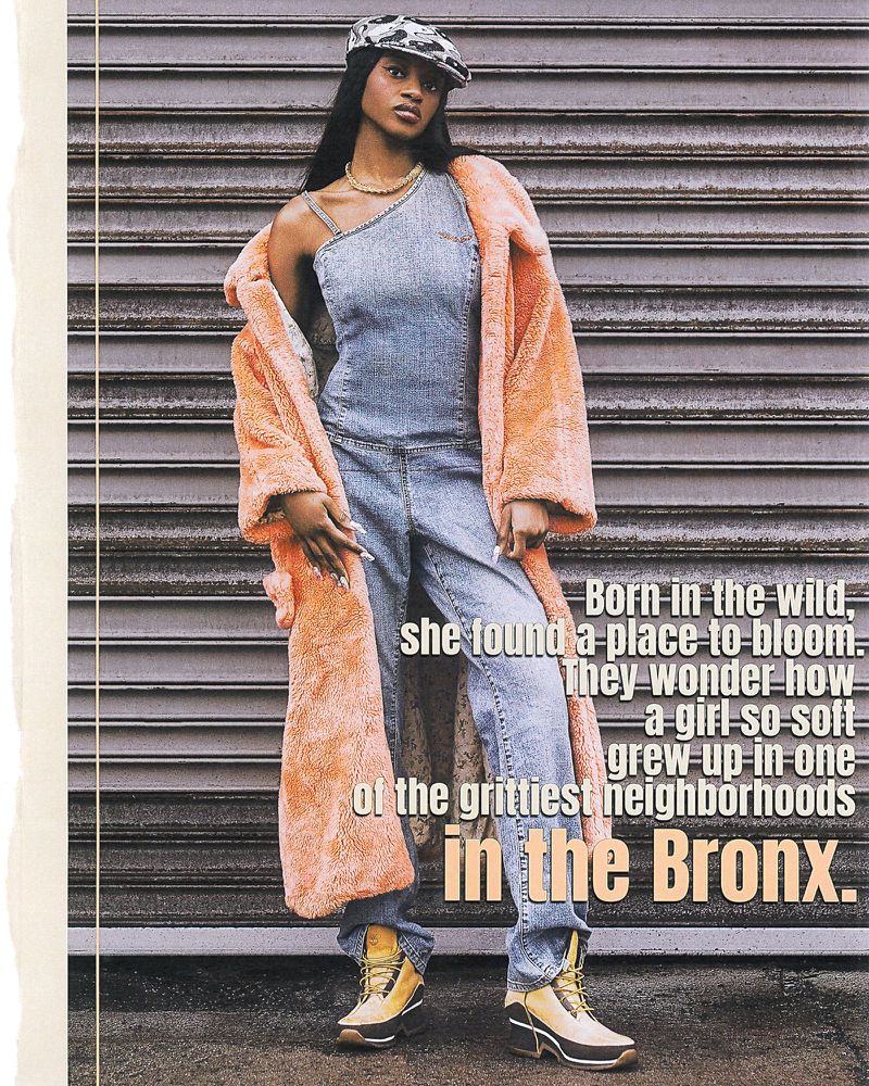 A young Black woman stands in front of a metal wall wearing a denim jumpsuit, peach coat, gold jewelry, and camouflage hat in a studio shot.