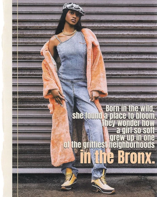 A young Black woman stands in front of a metal wall wearing a denim jumpsuit, peach coat, gold jewelry, and camouflage hat in a studio shot.