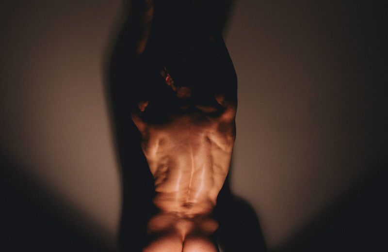 An athletic male figure with defined back muscles posing in a dimly lit indoor setting, creating dramatic contrasts and highlighting form and shadow.