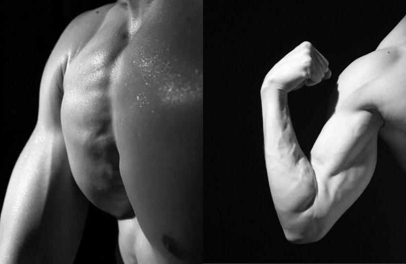 Close-up in black and white of the upper body of a muscular adult male with visible veins, muscles, and some sweat in a dark indoor setting.