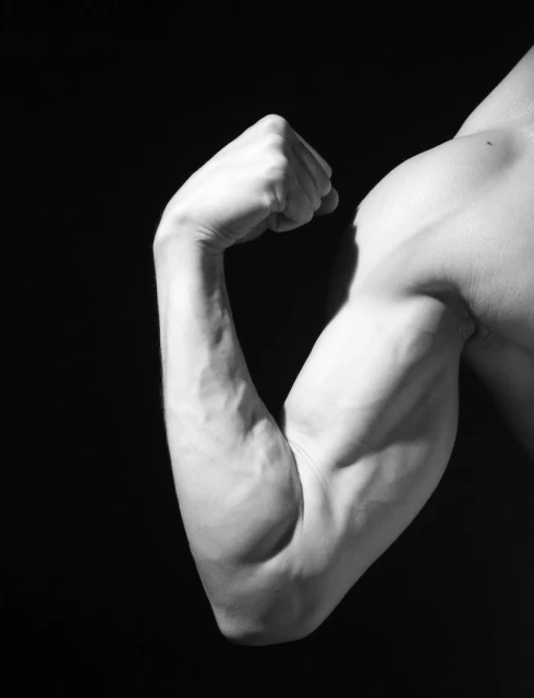 Monochrome shot of a muscular adult male flexing his biceps against a black backdrop, emphasizing fitness and strength with stark contrast.