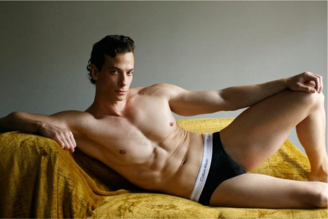 Athletic male model lying shirtless on a gold couch in a photo studio, wearing black Calvin Klein underwear and looking at the camera.