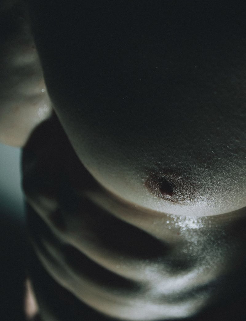 Close-up of an adult male torso exhibiting abdominal muscles, bathed in shadow and light, creating a dramatic aesthetic.