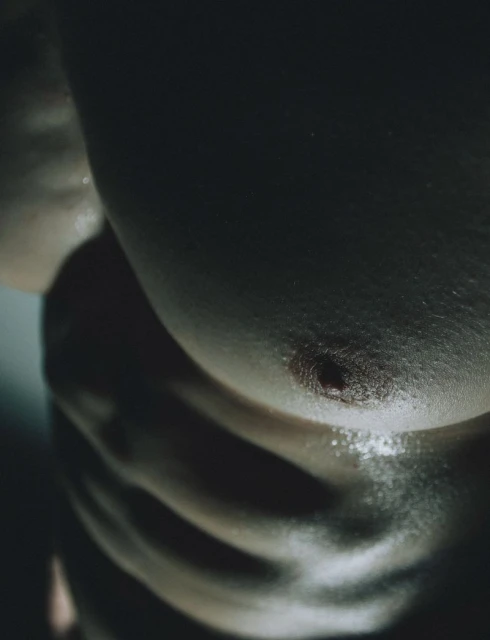 Close-up of an adult male torso exhibiting abdominal muscles, bathed in shadow and light, creating a dramatic aesthetic.