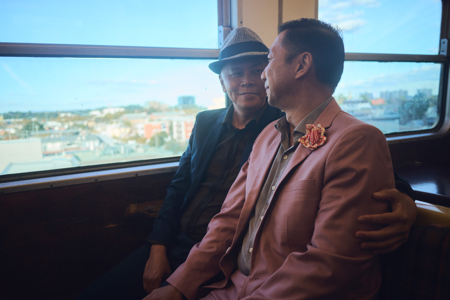 Two adult Asian men in a train car. One wears a grey hat and dark jacket, while the other wears a pink suit with a decorative lapel pin.
