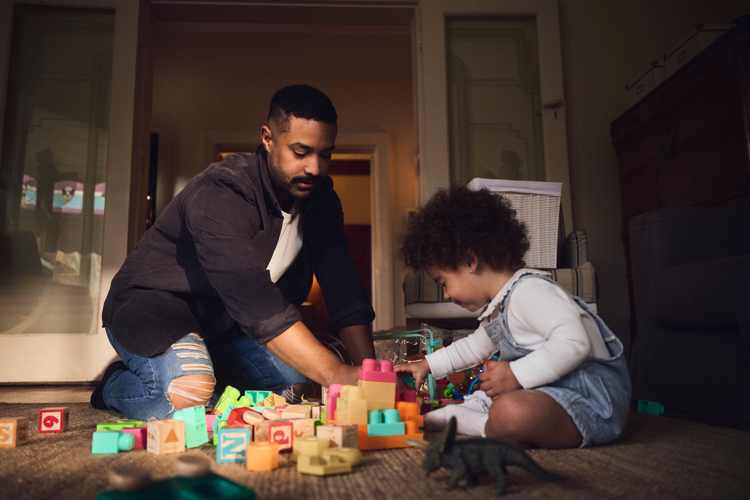 Father and toddler playing with building blocks on floor indoors. Dad assists young child with a tower of colorful plastic blocks.