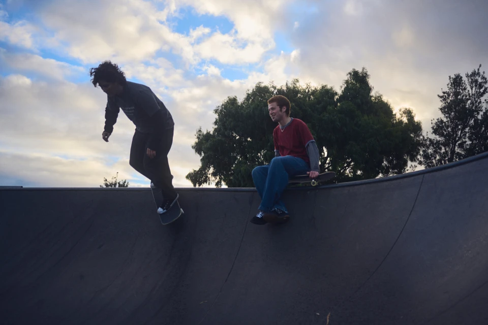 Two young men at a skatepark, one skateboarding down a ramp while the other sits at the edge of the ramp with his skateboard.