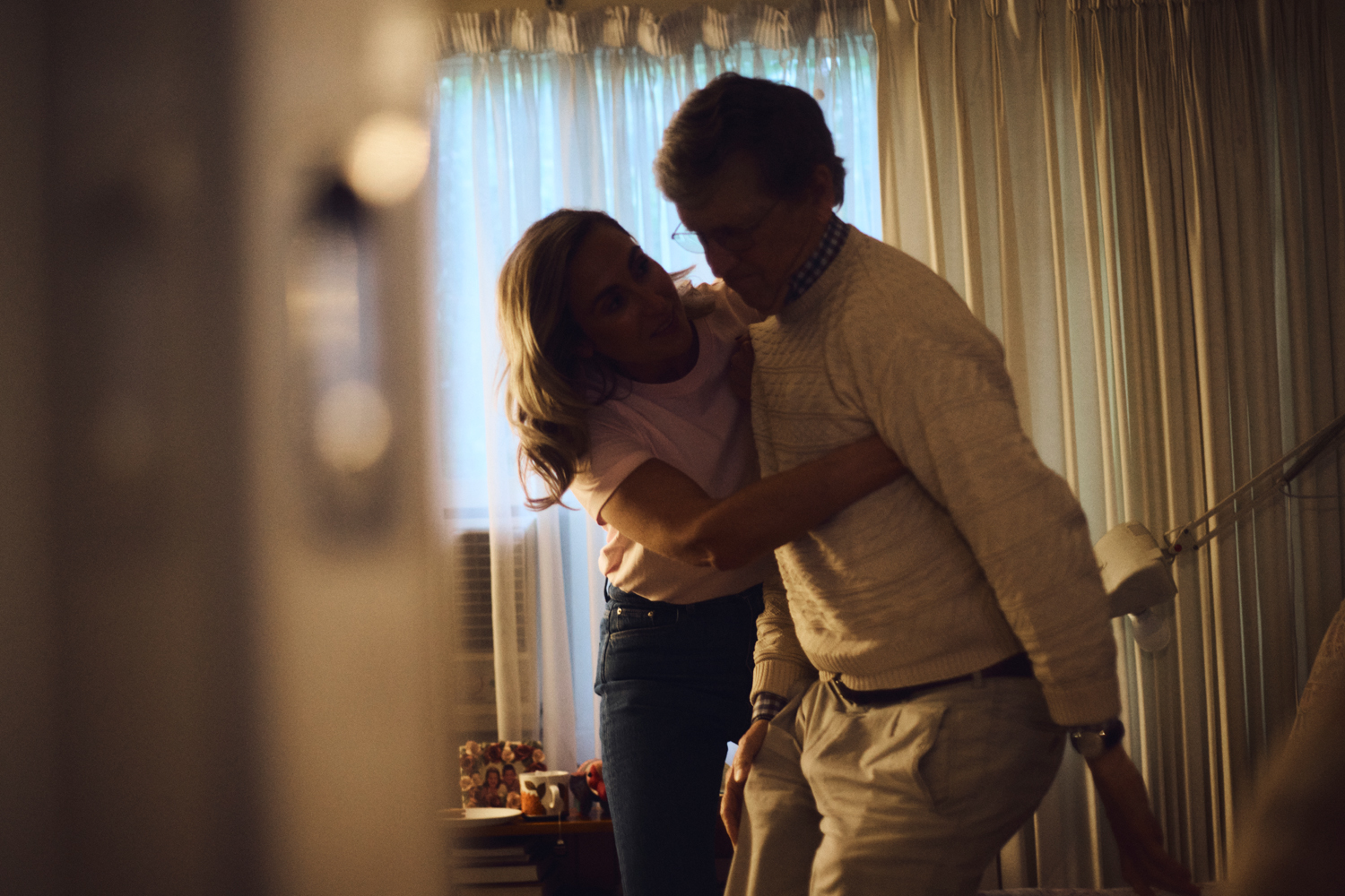 Woman embraces older man in warm indoor setting with sheer curtains, capturing a heartfelt moment with soft light and familial atmosphere.