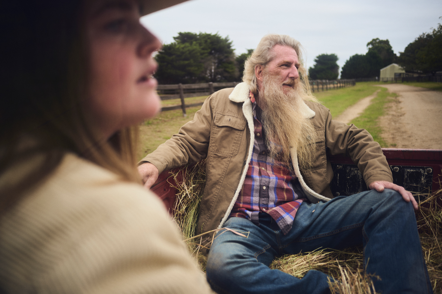 Mature man with long beard and young woman sit in truck bed on rural country road with trees and distant farmhouse in background.