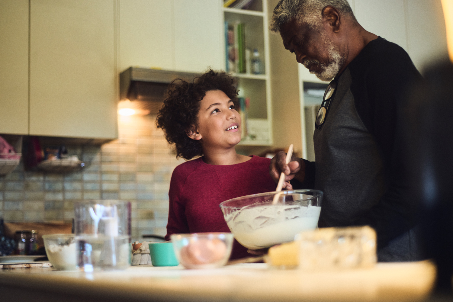 An elderly man and a young boy cooking together in a kitchen, mixing batter in a glass bowl.