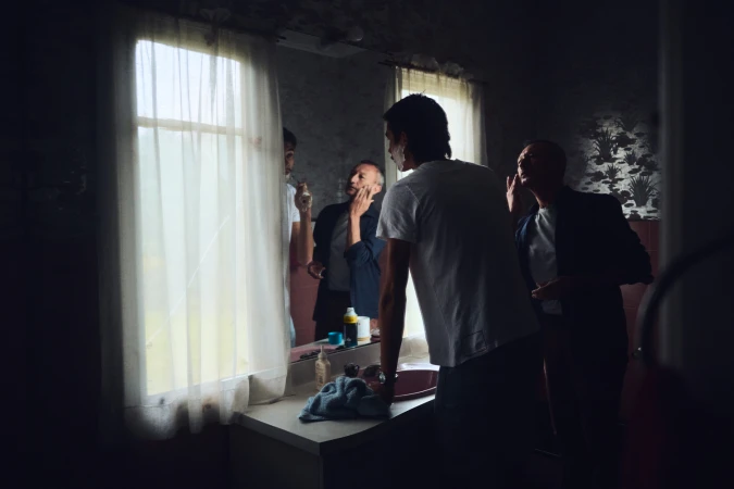 Three men standing in a domestic bathroom looking into a mirror by a window with sheer curtains