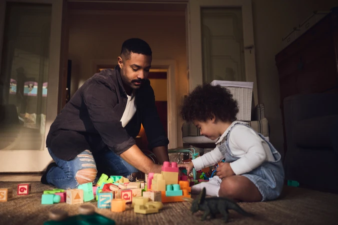 Man kneeling on floor playing with a child surrounded by colorful toy blocks in a living room