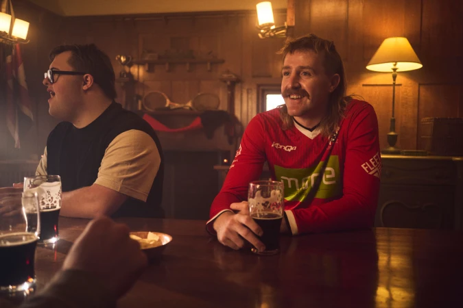 Two men sitting at a pub table with drinks, one wearing a red long-sleeve sports jersey, photographed by Seiya Taguchi for Movember.