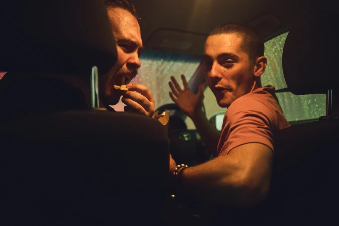 Two men inside a car at night, one eating fries in the driver seat and another looking back from the passenger seat.
