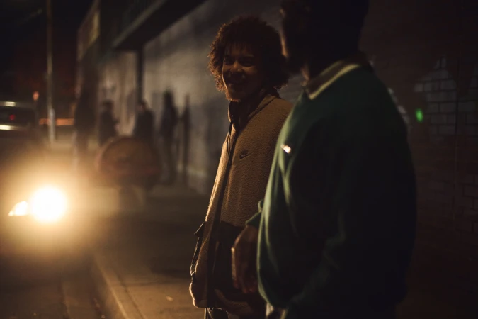 Two men standing outside at night on a dark city street next to a bright vehicle headlight and a brick wall